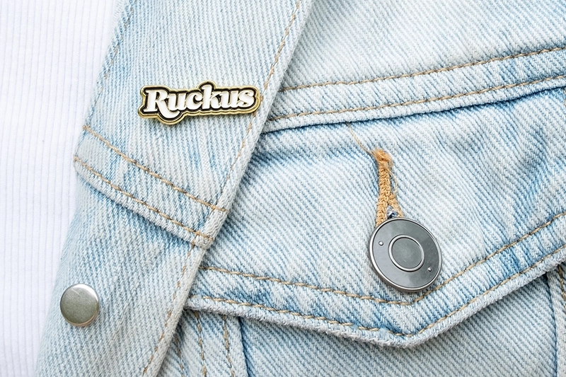 close up shot of a denim jacket with an enamel pin in the shape of the ruckus logo